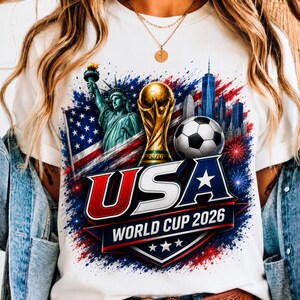 Puede incluir: Una camiseta blanca con un estampado gráfico de la Estatua de la Libertad, un trofeo de fútbol dorado, un balón de fútbol y un horizonte urbano. El diseño incluye salpicaduras de pintura roja, blanca y azul, fuegos artificiales y el texto USA.