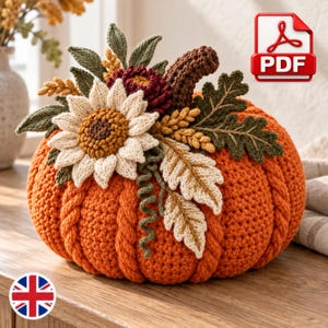 Pode incluir: Uma abóbora laranja de crochê com textura de tricô trançado, coberta por um caule marrom e um arranjo de girassóis creme, flores vermelho-escuras, folhas de carvalho verdes e espigas de trigo de crochê. Um ícone PDF vermelho é visível.