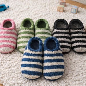 Può includere: Una collezione di pantofole a righe in vari colori, tra cui rosa, verde, nero, blu e beige. Le pantofole sono disposte su una superficie bianca e strutturata, con una piccola macchina giocattolo blu e dei cubi sullo sfondo.