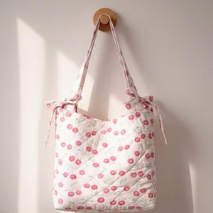 Könnte beinhalten: Eine gesteppte Tragetasche mit einem Muster aus rosa Kirschen und grünen Blättern auf weißem Grund. Die Tasche hat zwei mit Schleifen gebundene Henkel. Sie hängt an einem Holzpflock an einer weißen Wand.