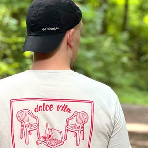 Könnte beinhalten: Eine schwarze Columbia Baseballkappe und ein weißes T-Shirt mit einem roten Grafikdruck auf der Rückseite. Der Druck zeigt zwei Plastikstühle, einen Pizzakarton, eine Flasche und einen Becher, mit dem Text "dolce vita" in roter Schreibschrift.