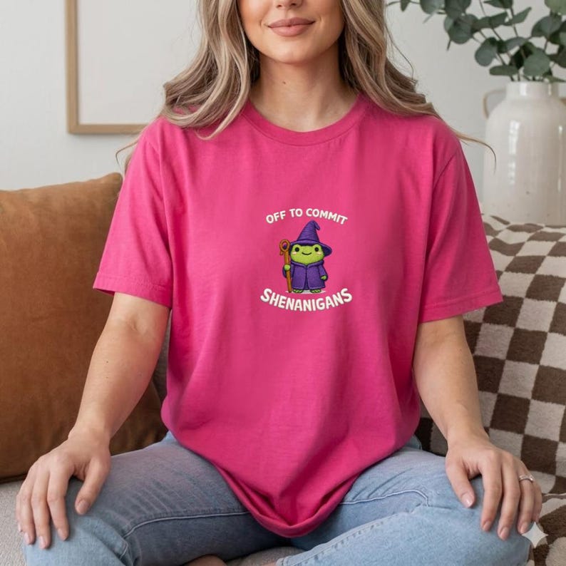 May include: A magenta t-shirt with the text "OFF TO COMMIT SHENANIGANS" and a cartoon frog wearing a wizard hat and robe. The frog is holding a staff. The shirt is a solid color and has short sleeves.
