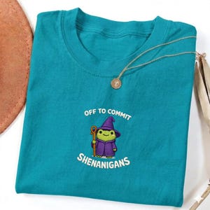 May include: Teal t-shirt with the phrase "OFF TO COMMIT SHENANIGANS" and a cartoon frog wearing a purple wizard hat and robe. A gold necklace with a pendant is draped across the shirt. The shirt is folded.