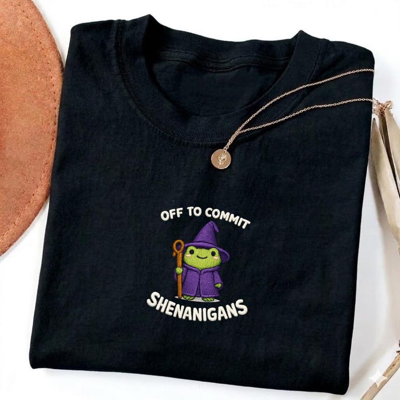 May include: Black t-shirt with the phrase "OFF TO COMMIT SHENANIGANS" and a cartoon frog wearing a purple wizard hat and robe. A gold necklace with a pendant is draped on the shirt.
