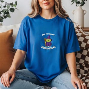 May include: Royal blue t-shirt with the text "OFF TO COMMIT SHENANIGANS" and a cartoon witch character. The witch is wearing a purple hat and cloak, holding a staff. The shirt is a crew neck style.