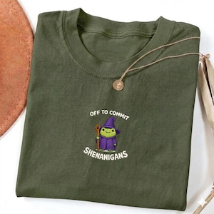 May include: An olive green t-shirt featuring the text "Off to Commit Shenanigans" and a cartoon frog in a wizard's hat. A gold-coloured necklace with a pendant is draped across the shirt.