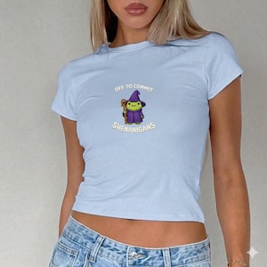 May include: Light blue t-shirt with a crew neck and short sleeves. The shirt features a cartoon graphic of a wizard frog with the text "OFF TO COMMIT SHENANIGANS" in a playful font. The frog is wearing a purple hat and robe.