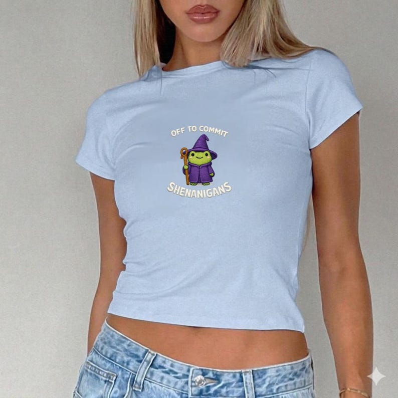 May include: Light blue t-shirt with a crew neck and short sleeves. The shirt features a cartoon graphic of a wizard frog with the text "OFF TO COMMIT SHENANIGANS" in a playful font. The frog is wearing a purple hat and robe.