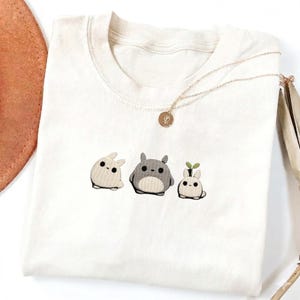May include: A white crew neck t-shirt featuring three embroidered cartoon characters. The characters are in shades of gray and white, with black dot eyes. A gold necklace with a small pendant is draped across the shirt.
