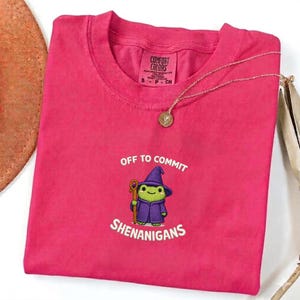 May include: A hot pink Comfort Colors t-shirt with the embroidered phrase "Off to Commit Shenanigans" and a cartoon wizard frog. A gold necklace with a round pendant rests on the shirt.