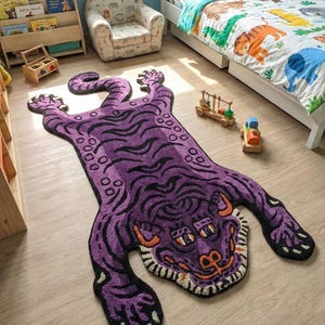 Op de afbeelding: Een paars tapijt in de vorm van een tijger met zwarte strepen en oranje en witte details. Het ligt op een lichte houten vloer in een kinderkamer, met een bed en speelgoed op de achtergrond. Lengte ongeveer 180 cm.