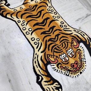Könnte beinhalten: Ein getufteter Teppich in Tigerform mit goldgelbem Körper, schwarzen Streifen und weißen Akzenten. Das Tigerdesign zeigt ein stilisiertes Gesicht mit zwei Augenpaaren, einer rosa Nase und einem geöffneten Maul mit Zähnen sowie einem cremefarbenen Bauch.