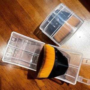 Op de afbeelding: Make-up kwastenset met een zwarte handgreep en oranje haren, gepresenteerd in een doorzichtige plastic doos. De set bevat meerdere kwasten, sommige met donkere haren. De doos is open.
