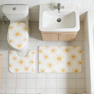 Könnte beinhalten: Ein Badezimmer-Set mit einem weißen Toilettensitzbezug, einer U-förmigen Badematte und einem rechteckigen Teppich, alle mit einem sich wiederholenden Muster aus gelben und weißen Gänseblümchen. Das Set befindet sich in einem Badezimmer mit weißen Fliesen.