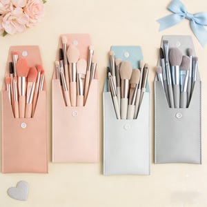 Op de afbeelding: Vier make-up kwastensets in pastelkleurige etuis. Elke etui heeft een andere kleur: roze, lichtroze, lichtblauw en grijs. De kwasten hebben lichtgeleurde handvatten en zachte borstelharen. Op de achtergrond een lichtblauwe strik en roze bloemen.