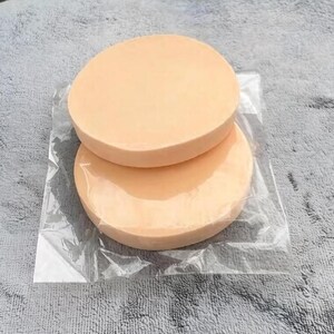 May include: Two peach-coloured, round makeup sponges are stacked inside a clear plastic package. The sponges have a smooth surface and are designed for applying cosmetics. The packaging is transparent, allowing the product to be visible.
