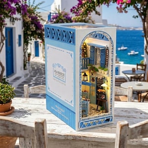 May include: A miniature, illuminated diorama of a Greek village scene. The model features blue and white buildings, a cobblestone street, and the text "SEA BREEZE". The diorama is set on a wooden table with a blurred background of a coastal landscape.