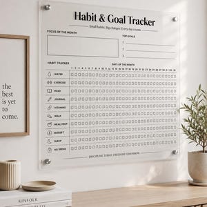 May include: A clear acrylic wall-mounted habit and goal tracker board with black printed text and icons. The board features sections for focus of the month, top goals, and a grid for tracking daily habits like water, exercise, and sleep.