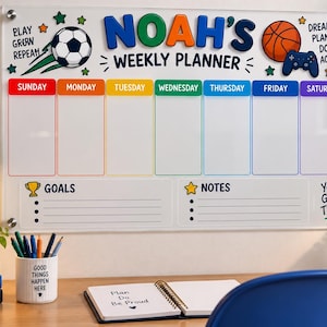 May include: A clear acrylic weekly planner board titled Noah's Weekly Planner with colorful headers for each day of the week. It features graphics of a soccer ball, basketball, and game controller, with sections for goals and notes, mounted with silver hardware.
