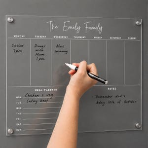 May include: A clear acrylic wall-mounted weekly calendar board with white printed text. The board features sections for a weekly schedule, a meal planner, and notes. It is titled The Emily Family and includes a black marker for writing tasks.