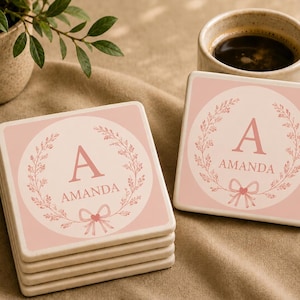 May include: A stack of square coasters and one individual coaster, each featuring a soft pink background with a white circular center. The design includes a dark pink letter A, the name AMANDA, a floral wreath, and a small bow at the bottom.