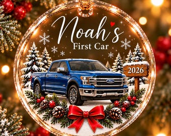 May include: A circular clear acrylic ornament featuring a blue Ford pickup truck, a wooden sign labeled 2026, and the text Noah's First Car. The design includes white snowflakes, green pine branches, red berries, and a red bow at the bottom.