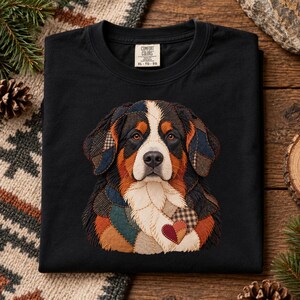 Puede incluir: Una camiseta negra con un gráfico de un perro boyero de Berna compuesto por varios parches de tela en tonos marrón, negro, blanco y azul. El diseño del perro incluye un pequeño parche en forma de corazón en el pecho. La camiseta tiene una etiqueta Comfort Colors.