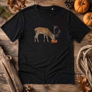 Può includere: Una t-shirt nera a maniche corte con un'illustrazione grafica di un cervo marrone con grandi corna che mangia una zucca arancione intagliata. Il capo presenta un girocollo e una vestibilità standard, con un design naturalistico dettagliato.