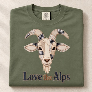 Puede incluir: Una camiseta verde oliva con un gráfico central de una cabeza de cabra hecha de varios parches de tela con patrones en azul, blanco y beige. Debajo de la cabra, el texto dice Love the Alps en letras bordadas de color azul oscuro y marrón.