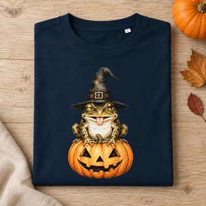 Può includere: Una t-shirt blu navy scuro con una stampa grafica di una rana che fa l'occhiolino, che indossa un cappello da strega nero con una fibbia dorata. La rana è seduta sopra una zucca arancione. La maglietta ha un'etichetta taglia XL visibile.