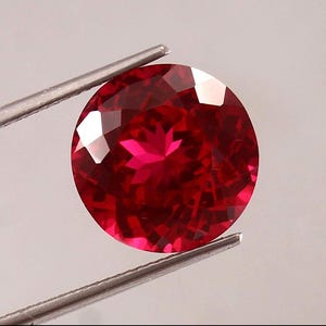 May include: A round, faceted, vibrant red gemstone held by silver-toned metal tweezers. The stone features a brilliant cut with multiple triangular facets that reflect light, creating a star-like pattern in the center of the deep red surface.
