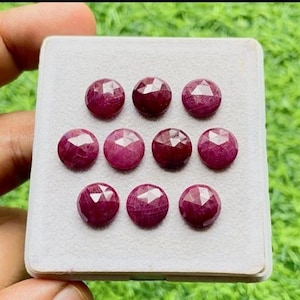 Könnte beinhalten: Eine Sammlung von zehn facettierten, runden, tiefroten Edelsteinen, möglicherweise Rubinen, wird auf einer weißen Oberfläche präsentiert. Die Edelsteine sind in drei Reihen angeordnet, jede mit einem einzigartigen Facettenmuster, vor einem Hintergrund aus grünem Gras.