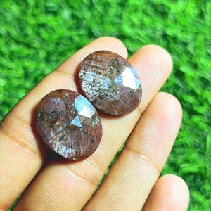 Könnte beinhalten: Zwei ovale, facettierte Edelsteine ​​mit rötlich-brauner Farbe werden in einer Hand gehalten. Die Edelsteine ​​haben eine polierte, reflektierende Oberfläche mit einem prominenten, vielseitigen Schliff. Der Hintergrund ist verschwommen grün.