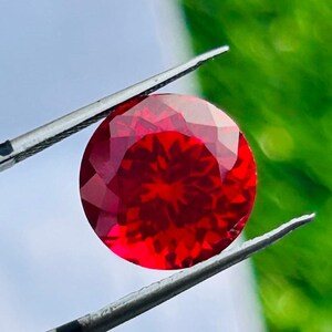 May include: A round, faceted, vibrant red gemstone held by metal tweezers. The stone features a complex, symmetrical geometric cut that reflects light, creating a star-like pattern within its deep crimson interior and polished, sharp edges.