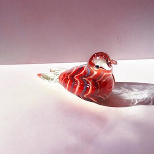 Puede incluir: Figura decorativa de pájaro de cristal. El pájaro es principalmente rojo con detalles de rayas blancas y azules. La cabeza es blanca con puntos rojos y un pico pequeño. Las alas son de cristal transparente. La figura está sobre una superficie blanca.