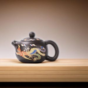 Peut inclure: Théière en céramique noire avec un motif de phénix décoratif rouge, jaune, bleu et vert. La théière a un corps arrondi, une anse incurvée et un petit bec. Le couvercle comporte un petit bouton et des nuages.