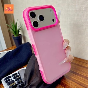 May include: A pink phone case with a bright pink border. The case has a matte finish and a camera cutout with a silver frame. The phone case is held by a person with long nails. The logo "Vy Crafter" is in the top left corner.