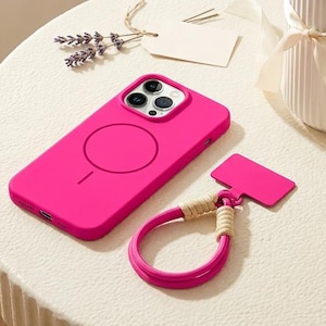 May include: A vibrant hot pink smartphone case featuring a circular MagSafe magnetic charging ring on the back, accompanied by a matching hot pink phone charm with a braided beige cord loop attachment, both displayed on a textured round surface.