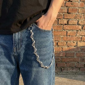 Op de afbeelding: Een zilverkleurige metalen portemonneeketting met een gestructureerd, slangachtig schakelontwerp. De ketting is bevestigd aan de riemlus van een blauwe spijkerbroek en hangt in een gebogen, decoratieve vorm over het voorste zakgedeelte.