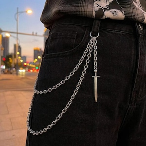 May include: A silver-toned metal wallet chain attached to black denim jeans. The accessory features two layered chain strands and a small sword-shaped pendant charm hanging from the belt loop attachment point on the side of the garment.