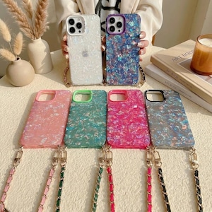 May include: A collection of six smartphone cases featuring a shimmering, iridescent shell-like pattern. Each case includes a matching chain strap with gold-tone hardware. The colours include white, blue, pink, green, magenta, and dark grey, all with cutouts.