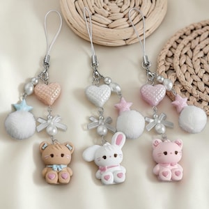 May include: Three decorative phone charms with silver-tone hardware. Each charm features a small animal figurine, a heart, a star, a fluffy ball, and a bow. The animal figurines include a bear, a bunny, and a cat, all in pastel colors.