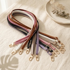 May include: A collection of adjustable bag straps in various colors including brown, white, pink, black, gray, and purple. Each strap features a textured surface, gold-toned metal buckles, and gold-toned lobster claw clasps at both ends.
