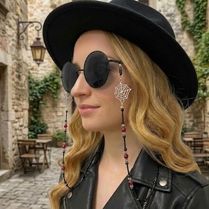 Puede incluir: Un sombrero fedora negro, gafas de sol redondas negras y una cadena decorativa para gafas con dijes plateados de telaraña, pequeñas cuentas negras y cuentas de color rojo oscuro unidas a las monturas de las gafas.