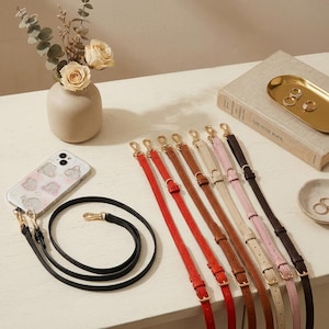 Könnte beinhalten: Eine Sammlung verstellbarer Taschenriemen in Rot, Braun, Hellbraun, Beige, Rosa und Schwarz mit goldfarbenen Metallbeschlägen und Karabinerhaken. Eine transparente Handyhülle mit rosa-grauem Muster ist an einem schwarzen Riemen befestigt.