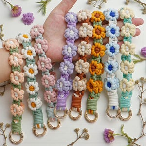 May include: A selection of handmade macramé keychains in various colours, including pink, white, yellow, blue, and purple. Each keychain features a daisy flower design and a gold-coloured metal ring. The keychains are held in a hand.