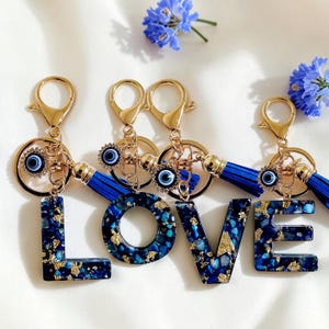 May include: Gold keyrings spelling "LOVE" with blue and gold detailing. Each letter is filled with blue stones and gold flakes. The keyrings have blue tassels and evil eye charms, with a lobster clasp for attachment.