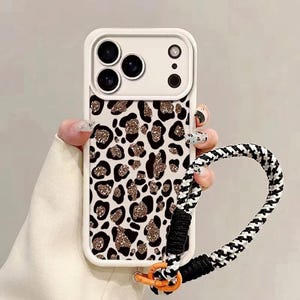 Può includere: Una custodia per smartphone bianca con un motivo leopardato nero e oro glitterato. La custodia include una cornice rialzata per la fotocamera e un cinturino da polso intrecciato rimovibile con motivo pied-de-poule bianco e nero e connettore arancione.