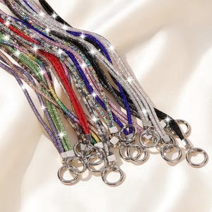 May include: A collection of lanyards featuring sparkling rhinestone-encrusted straps in various colors including red, blue, green, purple, black, and silver. Each strap is finished with a silver-toned metal circular ring clasp at the end.