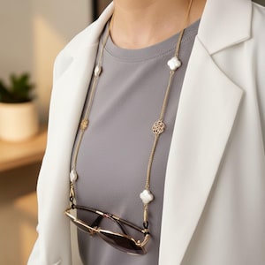 Op de afbeelding: Goudkleurige brillenketting met witte klaver- en bloemaccenten. De ketting houdt een zonnebril met bruine glazen vast. De ketting wordt gedragen over een grijs shirt en een witte blazer.